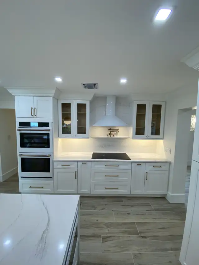 White kitchen with shaker cabinets, lit glass-front uppers, a white range hood, double wall ovens and wood-look plank tile