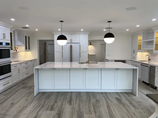 Renovated kitchen with a large white waterfall-edge quartz island, white shaker cabinets, black and white globe pendants and wood-look plank tile floor